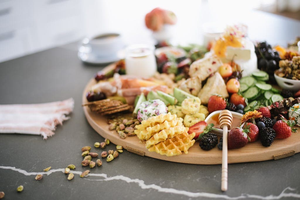 Wooden tray with mixed berries and honey with dipper served with waffles and sweets near cucumber slices on table in light room