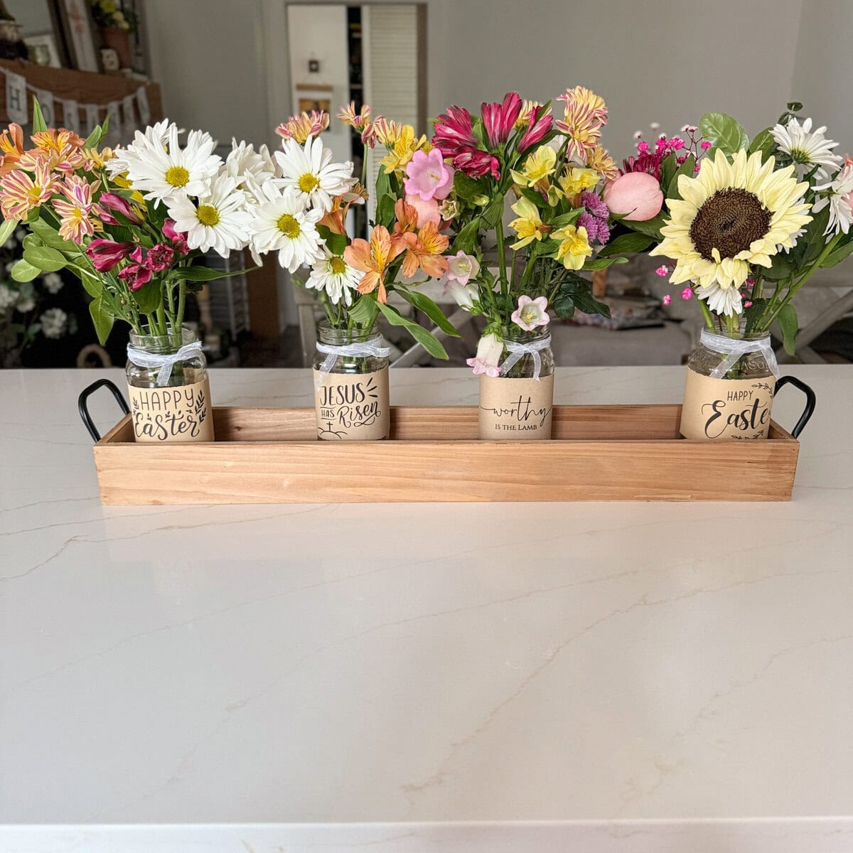 Vase floral arrangements with spring flowers in vintage mason jars decorated with Easter and religious-themed handwritten tags, presented in a wooden tray on a clean white countertop.
