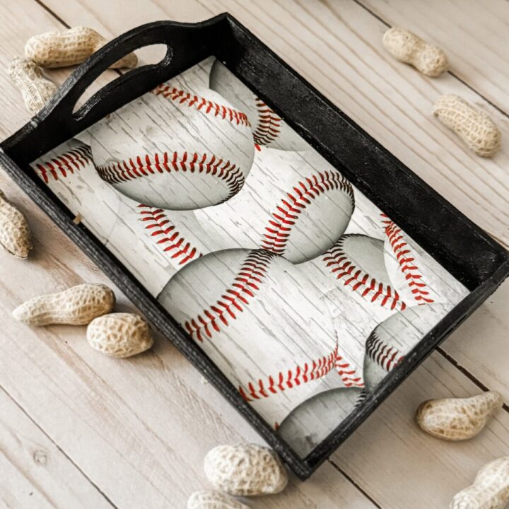 Vintage baseballs with red stitching in a rustic black wooden tray. Perfect for sports-themed decor or baseball enthusiasts, displayed on a light wooden surface with peanut shells around.