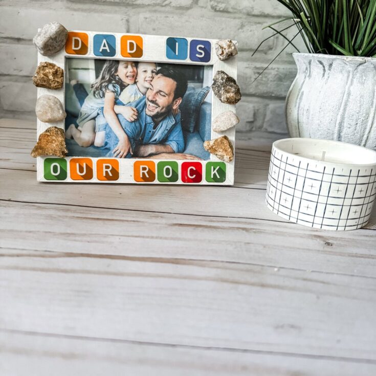 Happy family photo frame with "Dad is our rock" message, surrounded by decorative stones, on a rustic wooden surface for Father's Day or family celebration.
