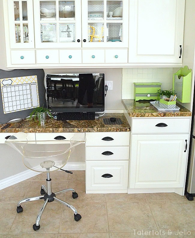 Aesthetic home office workspace in a bright, organized kitchen nook with white cabinetry, a granite countertop, a computer monitor, and colorful storage boxes for a functional and stylish environment.