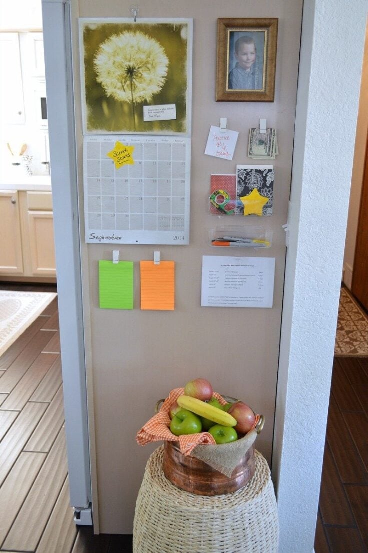Colorful kitchen calendar and family photos on a beige wall, with a basket of fresh apples, bananas, and peaches on a woven stand. A cozy, organized space promoting family life and healthy living.