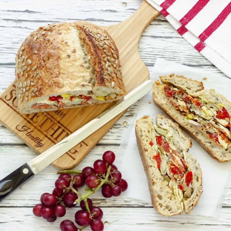Fresh baked vegetable and cheese stuffed bread with sunflower seeds, served on a rustic wooden cutting board with a bread knife, accompanied by a bunch of red grapes. Perfect for healthy snacking or light lunch.
