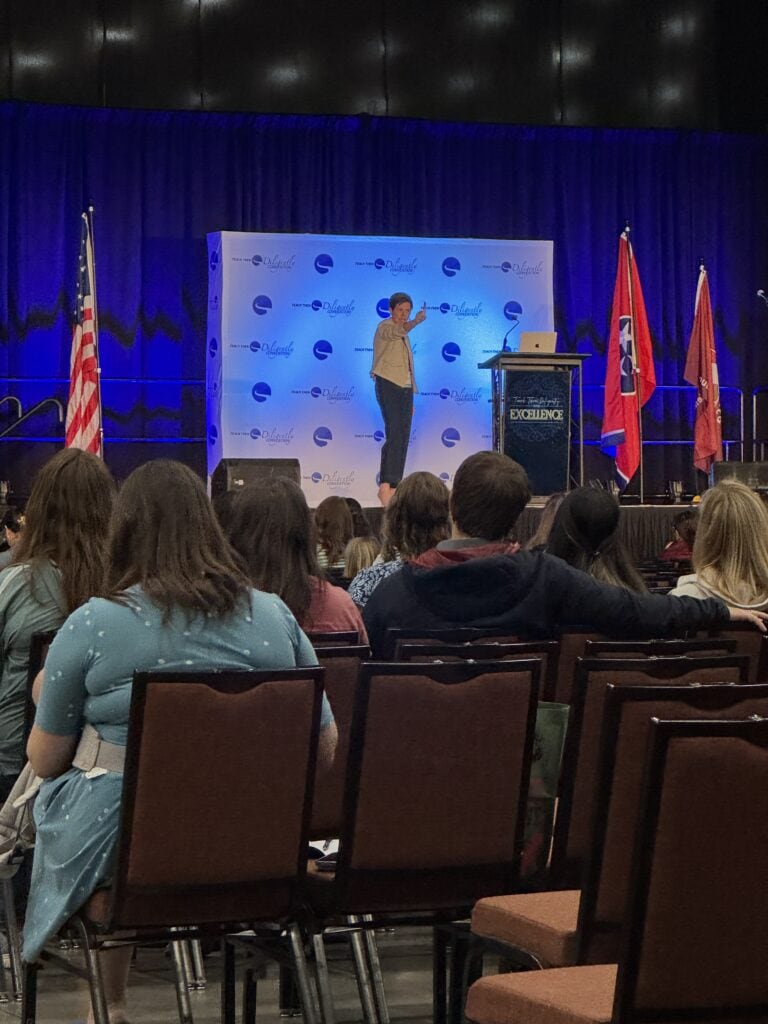 Empowering woman speaker on stage at a conference, addressing an attentive audience with flags and a branded backdrop, highlighting leadership, motivation, and professional development.