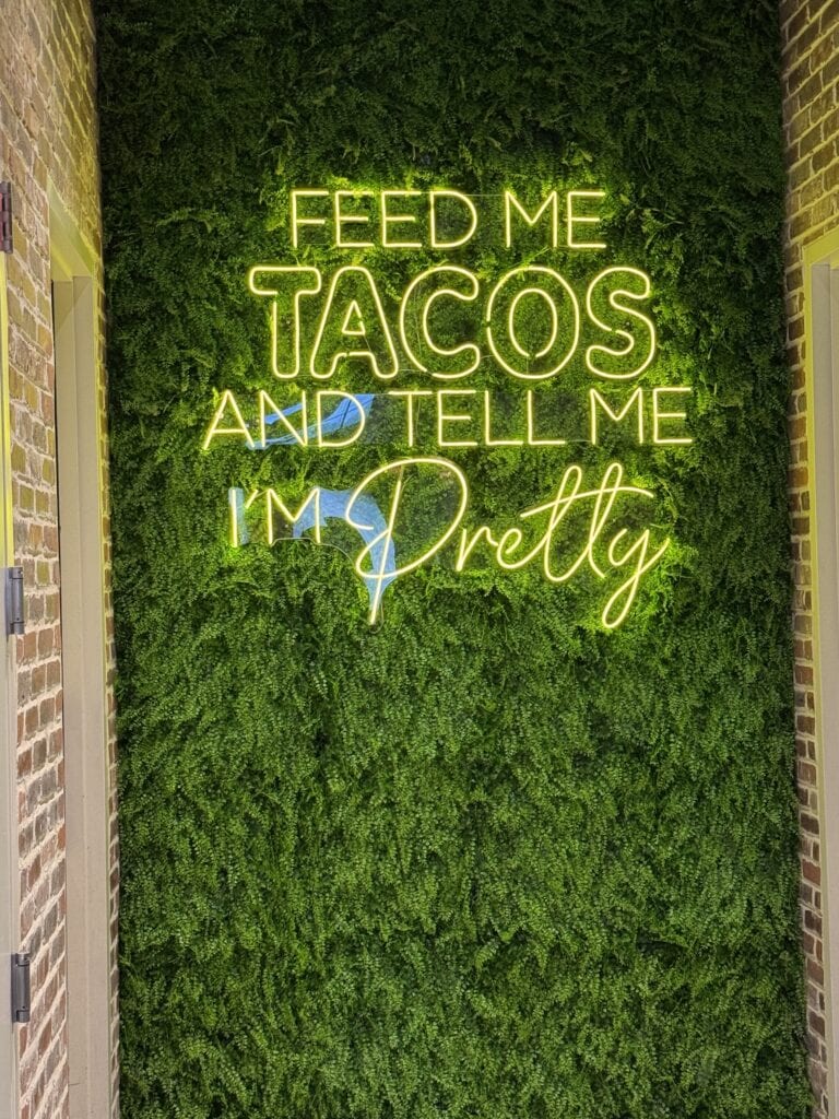Feed me tacos and tell me I'm pretty neon sign on a green moss wall at A Hundred Affections.