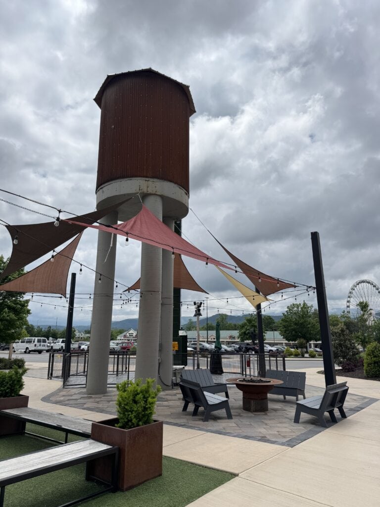 Vintage water tower with colorful shade sails at A Hundred Affections outdoor seating area, scenic view with cloudy sky and nearby Ferris wheel, featuring modern design and cozy ambiance.