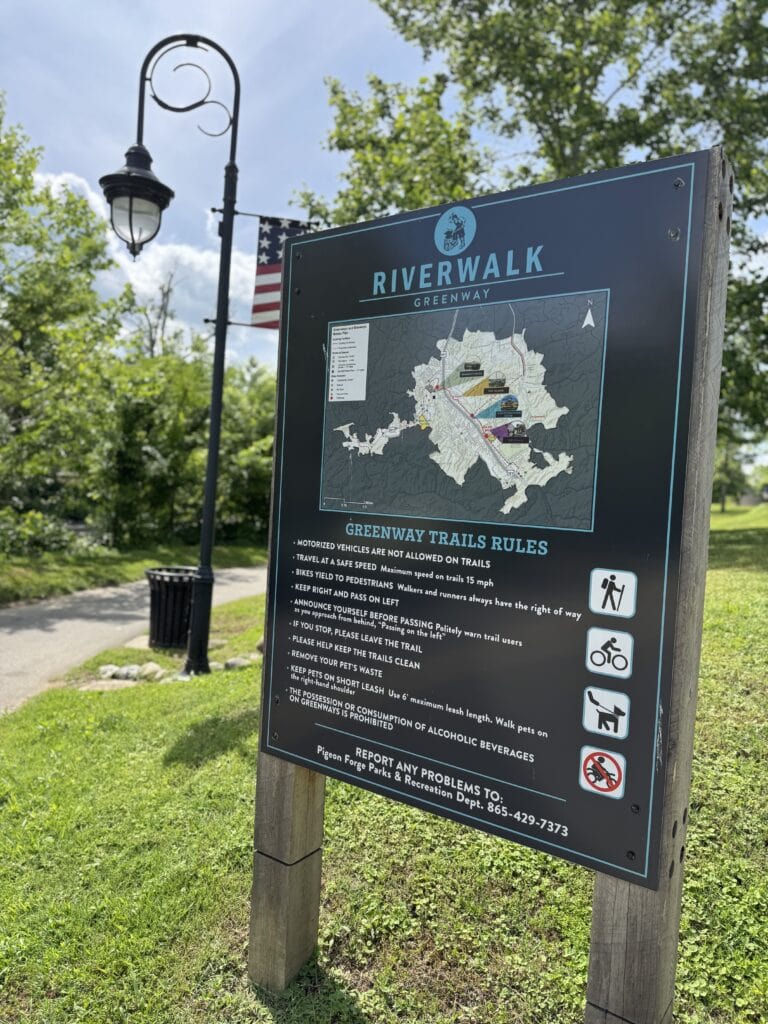 Colorful Riverwalk Greenway trail map with park rules and icons, set in a lush park with trees, a vintage street lamp, and an American flag, illustrating outdoor recreational activities and community space.