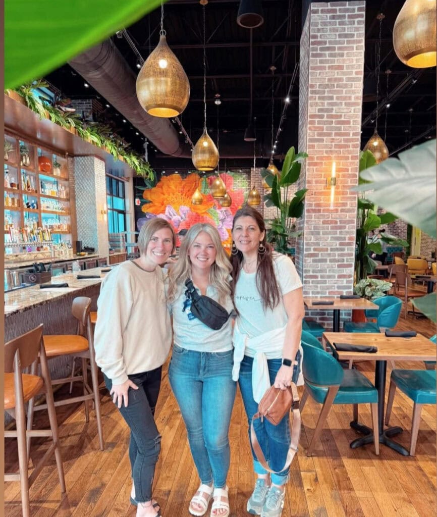 Beautiful women enjoying a vibrant, cozy cafe with lush greenery, modern lighting, and colorful floral wall art, perfect for socializing and capturing memorable moments.