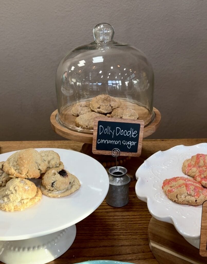 Butter cookies on a stand with a glass dome, labeled "Dolly Doodle cinnamon sugar" at A Hundred Affections bakery.