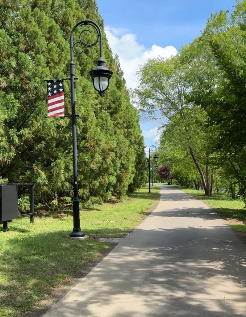 A peaceful park pathway with vintage-style black lampposts decorated with American flags, surrounded by lush green trees and vibrant foliage, under a bright blue sky.