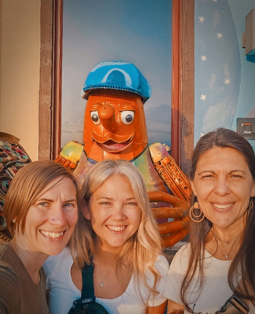 Vibrant photo of three women smiling in front of a colorful, whimsical statue with a whimsical face and pirate hat, perfect for family travel or fun adventures.