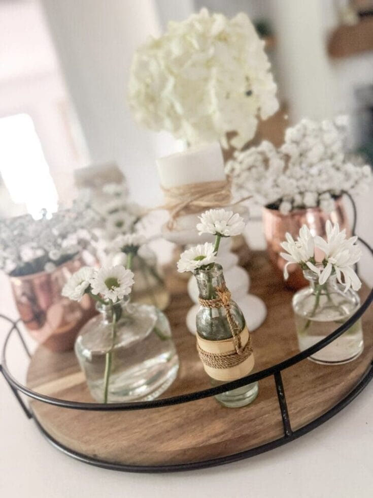 Soft white flowers in small glass vases, decorative candles, and copper accents on a round wooden and metal tray, perfect for home decor or floral arrangements.