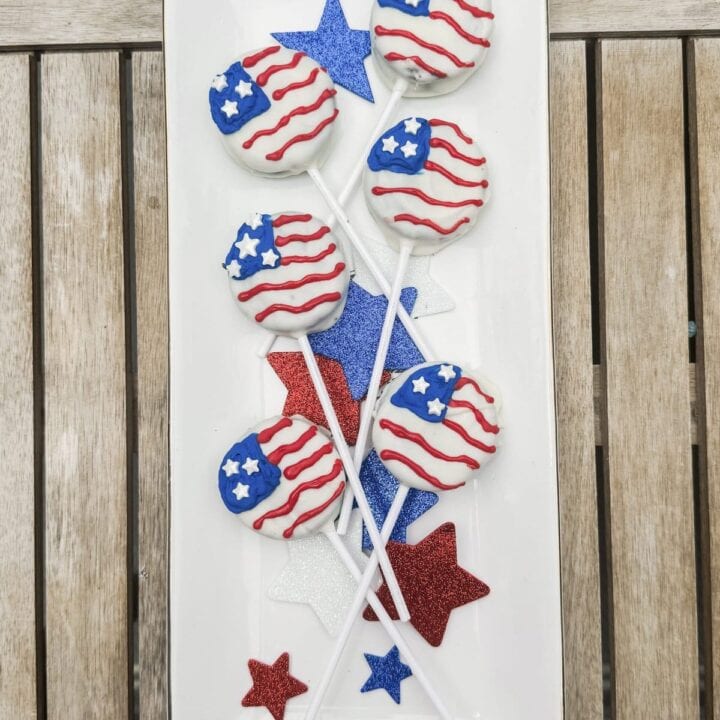 Patriotic American flag-themed cake pops decorated with red, white, and blue icing, stars, and stripes, perfect for Fourth of July celebrations, displayed on a white rectangular platter with red, white, and blue star decorations.