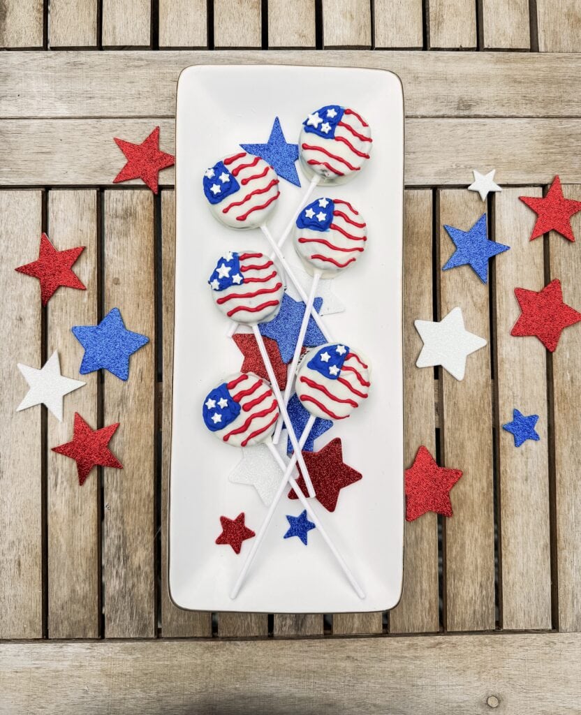 Red, white, and blue patriotic cake pops with American flag design, on a white rectangular platter, surrounded by red, white, and blue star-shaped confetti on a wooden surface.