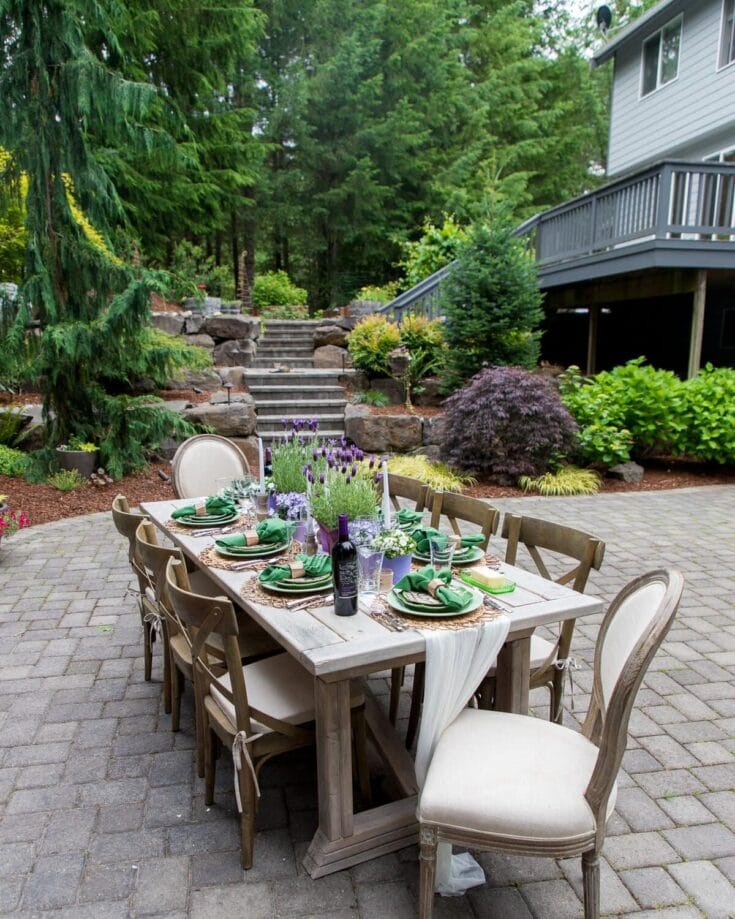 Outdoor garden dining table decorated for a special occasion with green napkins, wine bottles, and floral arrangements, set in a lush backyard with greenery and stone steps.
