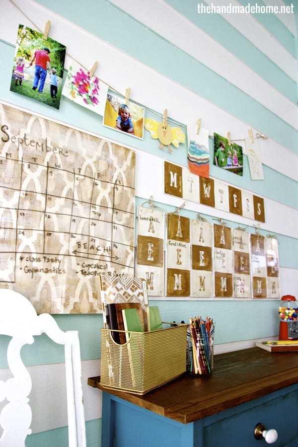 Photo of a colorful home hallway decorated with family photos, a monthly calendar, and a craft station on a blue wall with a wooden desk and storage bins.