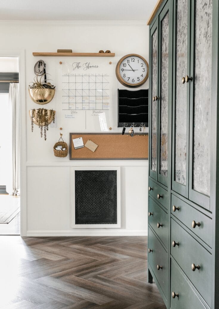 A cozy entryway with a white wall featuring a chalkboard calendar, two gold baskets hanging on hooks, and a black wall organizer; a green wooden cabinet with distressed finish adds rustic charm.