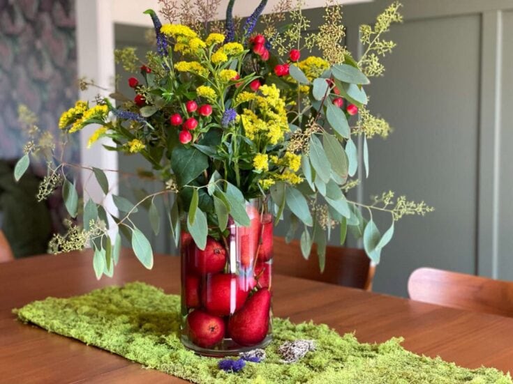 Red apples and vibrant wildflowers arranged in a tall glass vase on a wooden table, creating a fresh and lively centerpiece for home décor and floral arrangements.