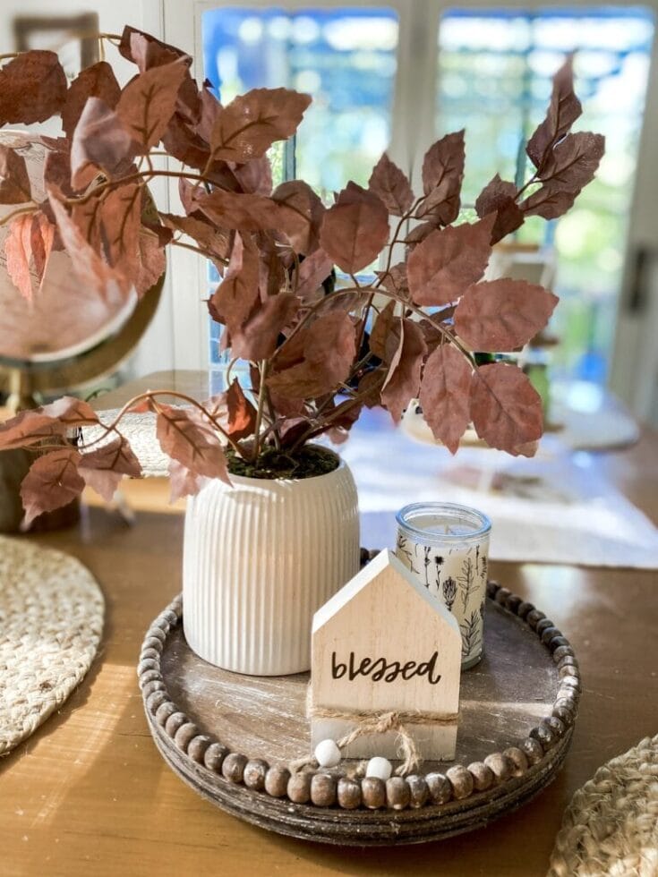 Rust-colored potted plant with dried leaves on a wooden tray with decorative "blessed" sign, candles, and cozy home decor, creating a warm, autumn-inspired centerpiece for home or gifts.