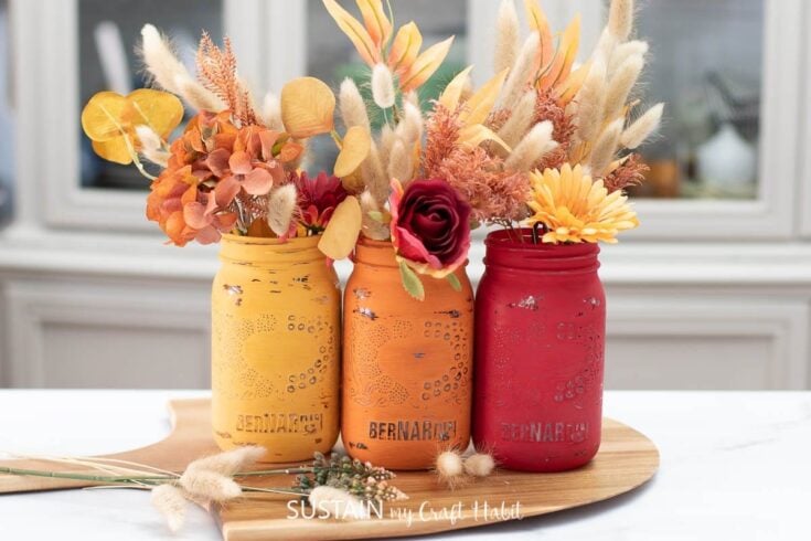 Vibrant fall floral arrangements in colorful painted Mason jars on a wooden tray, perfect for autumn decor and craft projects.