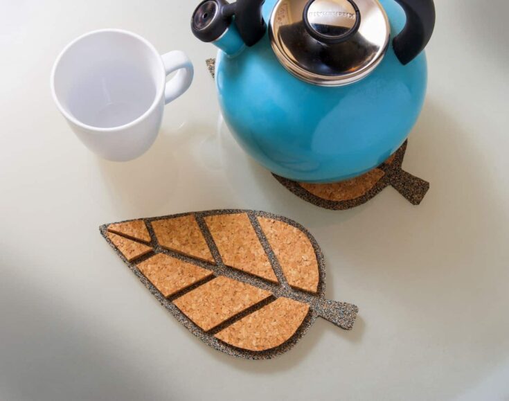 Colorful blue kettle with black handle and silver lid alongside a white coffee mug on a cork leaf-shaped coaster, creating a cozy, aesthetic kitchen scene.