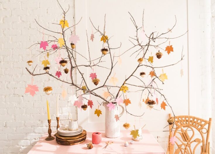 Vibrant fall-themed table centerpiece with a branch arranged in a white vase, decorated with colorful paper leaves, pinecones, and seasonal accents for cozy autumn gatherings.