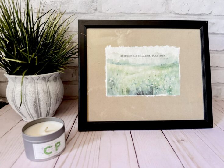 Soft watercolor painting of a lush green field with flowers, paired with a framed inspirational quote about creation, positioned on a pink wooden table with a white potted plant and candle for a serene, faith-inspired decor, perfect for religious or spiritual home accents.