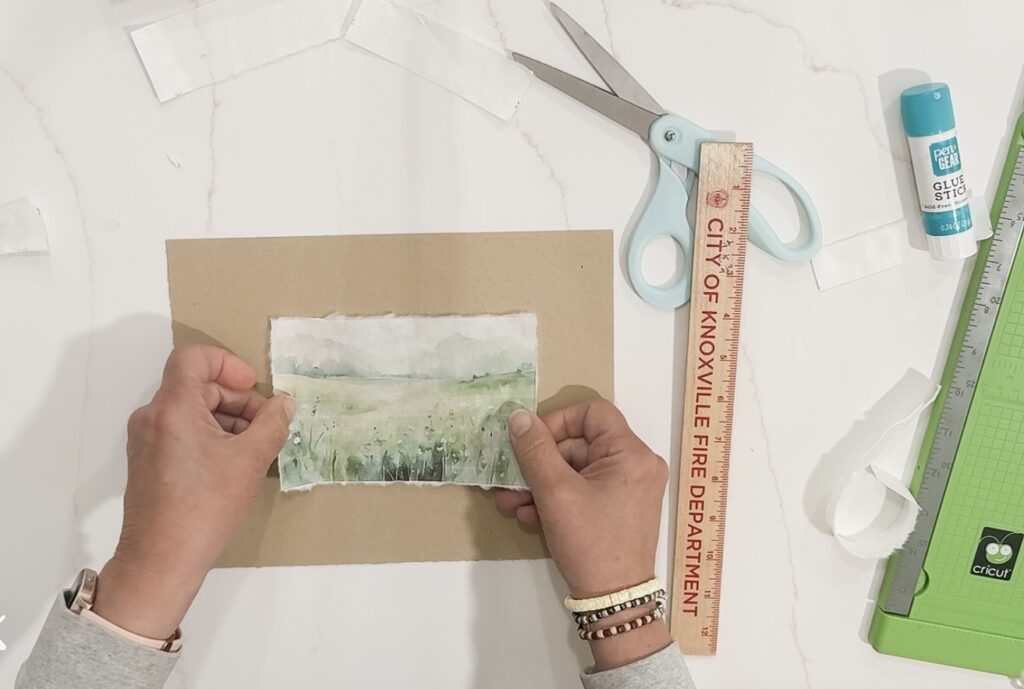 Delicate watercolor landscape art in progress, hand-held by artist with crafting tools and supplies on a white workspace, emphasizing handmade nature and artistic creativity.