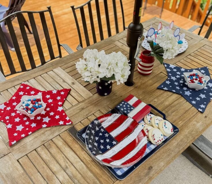 Patriotic Fourth of July table decor with American flag-themed plates, cookies, and candy, complemented by white hydrangea flowers in star-spangled vases, set on a rustic wooden outdoor table.