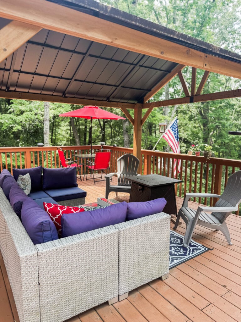 Colorful outdoor deck with cozy seating, red patio umbrella, American flag, surrounded by lush greenery, perfect for home relaxation and outdoor living.