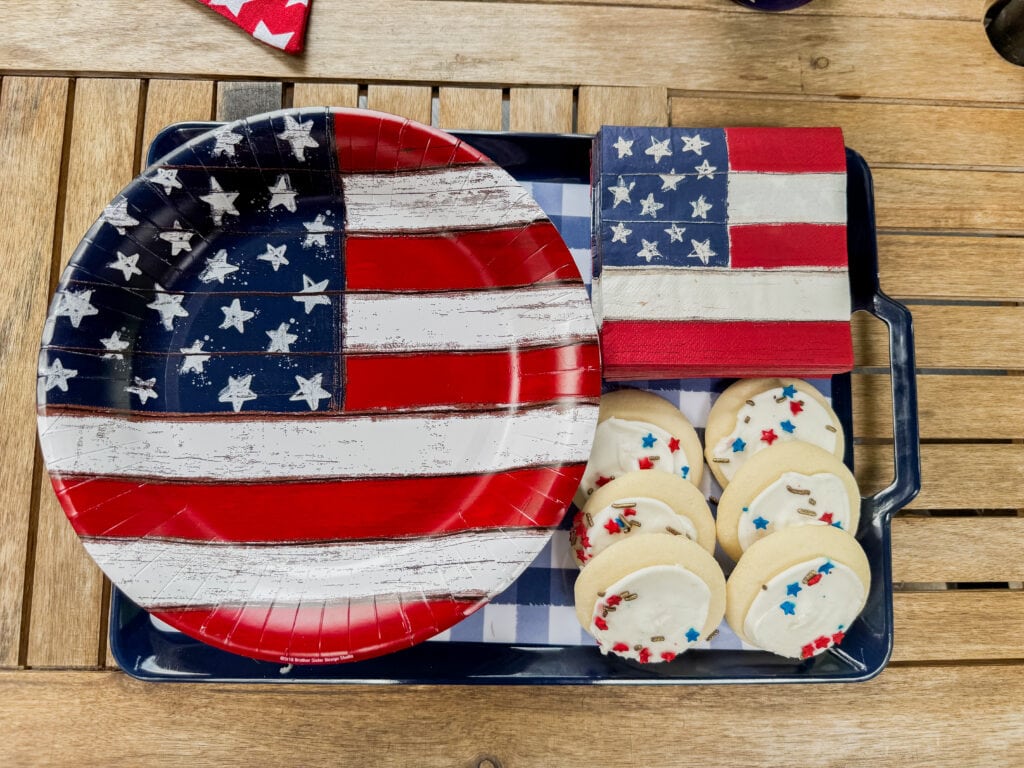 Red, white, and blue American flag-themed serving tray with patriotic paper napkin, plate, and cookies with stars and sprinkles, placed on a wooden table.