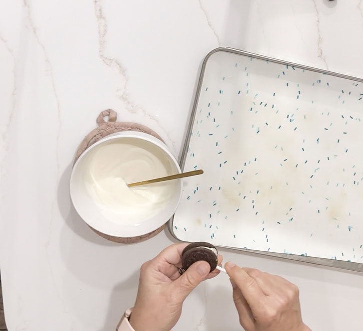 Cream cheese mixture in bowl with spoon, person's hands holding Oreo cookie, baking tray with blue sprinkles on marble surface, dessert preparation, sweet treat, baking, snack, dessert recipe, A Hundred Affections.