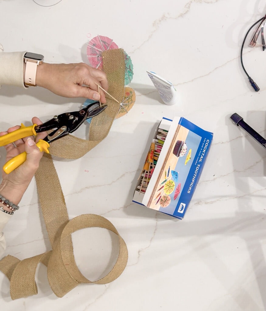 Colorful cocktail umbrella craft project on a white marble table, creating decorative accessories for summer parties or tropical-themed events, utilizing scissors, burlap ribbon, and assorted tools.