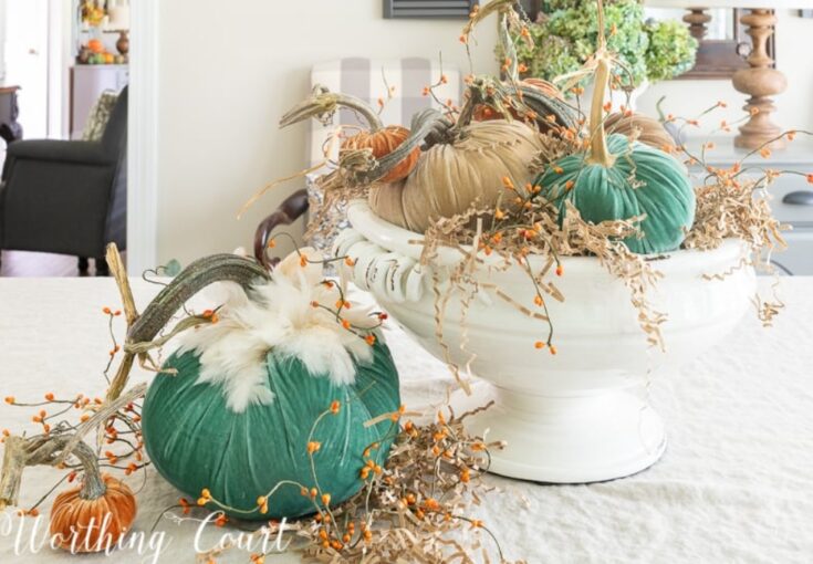 Colorful velvet pumpkins with faux autumn leaves and twigs arranged in a decorative white bowl for fall home decor.