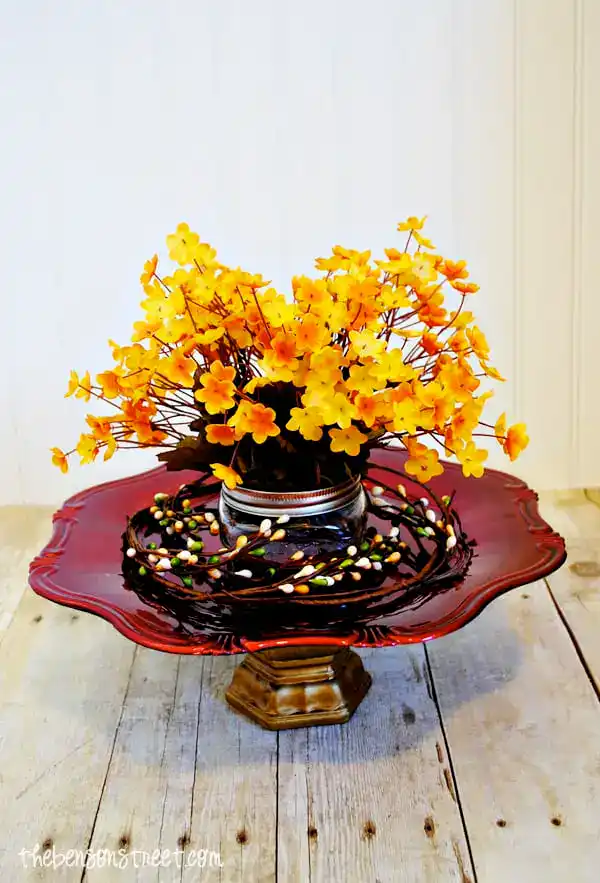 Vibrant yellow artificial flower arrangement in a silver pot, displayed on an elegant red wooden pedestal table with decorative Easter eggs, perfect for spring home decor.