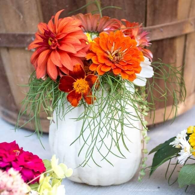 Vase with vibrant orange and red dahlias, marigolds, and greenery in a rustic white ceramic container, perfect for floral decor and creative arrangements.