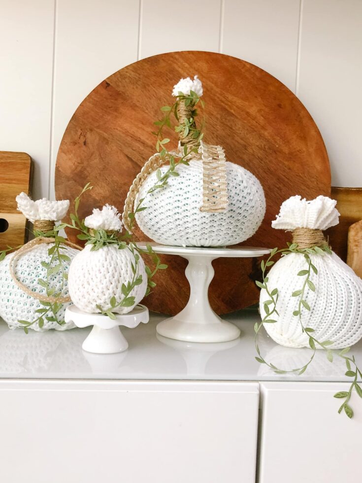 Handmade crocheted pumpkin decor with greenery, perfect for rustic or farmhouse seasonal decor, displayed on white cake stands with a wooden backdrop.