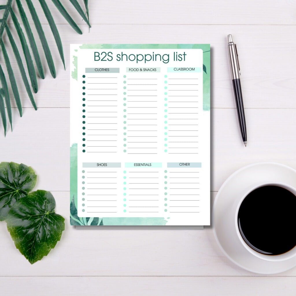 Organized B2S shopping list template with categories for clothes, food, classroom supplies, shoes, essentials, and others, on a white wooden desk with a black pen, coffee, and green plants.