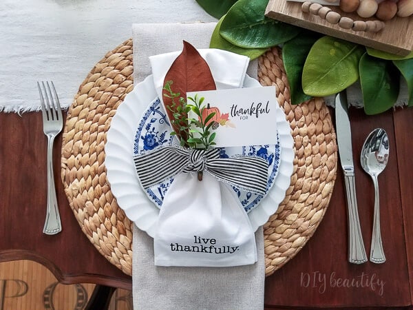 Beautiful table setting with a white napkin tied with a striped ribbon, a thankful card, and a greenery sprig, perfect for a gratitude-themed celebration or Thanksgiving dinner.