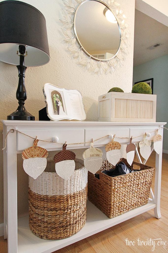 Soft white console table decorated with basket storage, a black table lamp, and a photo album, with a round mirror and decorative moss balls on the wall, creating a cozy, charming entryway or living space atmosphere.
