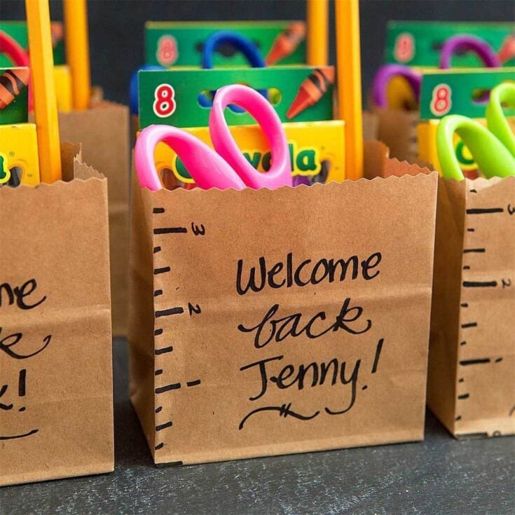 Colorful children's scissors and bubble gum in paper goodie bags with "Welcome back Jenny!" handwritten on the front, perfect for back-to-school or classroom welcome messages, cheerful and playful.