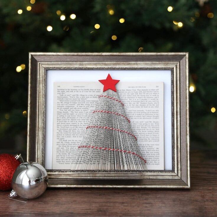 Handmade Christmas tree art framed with book pages, featuring a star on top and red string garland, displayed on a wooden surface with festive ornaments and blurred holiday lights in the background.