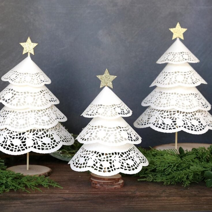Delicate white paper Christmas trees with intricate lace patterns, topped with glittery gold stars, arranged on a rustic wooden surface with greenery, perfect for holiday home decor and festive decorating.