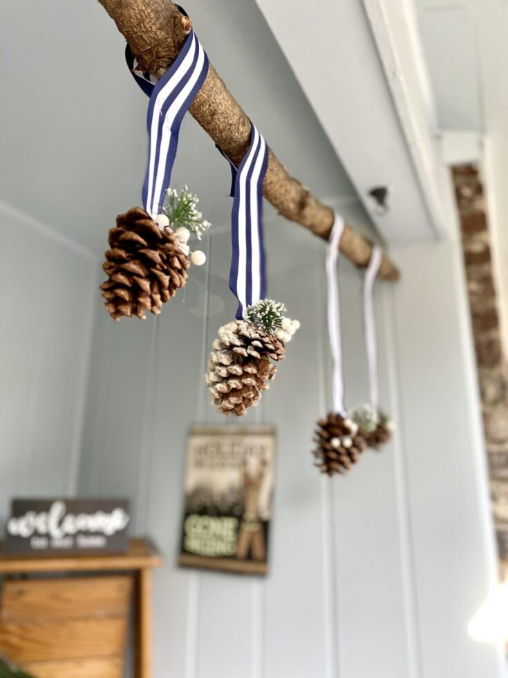 Pinecone holiday ornaments hanging from a rustic wooden branch with white and navy striped ribbons, decorated with small white berries and greenery, creating a cozy winter decor.