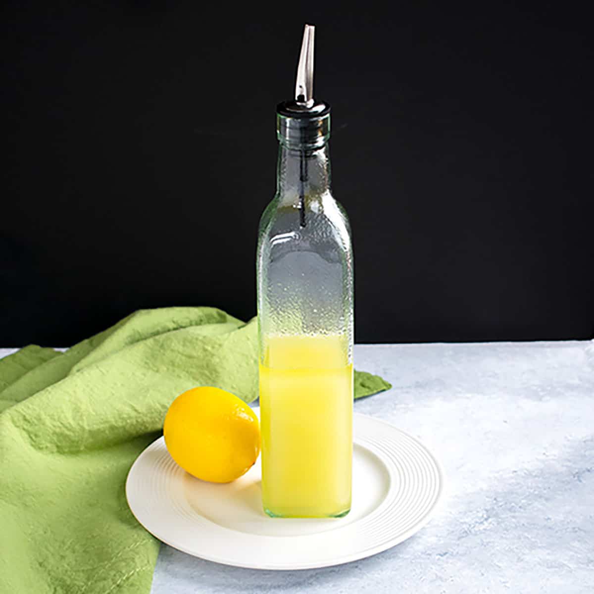 Refreshing homemade lemonade in a glass bottle with a metal pour spout, alongside a whole lemon, on a white plate with a green cloth napkin, on a textured surface against a black background.