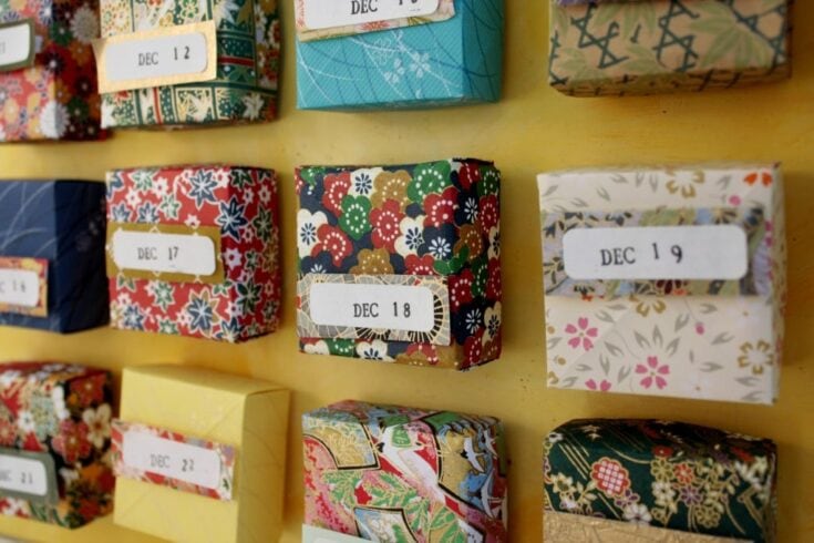 Colorful handmade advent calendars wrapped in decorative paper with December date labels, displayed on a yellow background, showcasing craft holiday gift ideas and festive decorations.