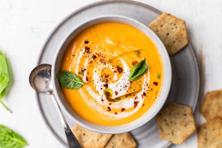 Creamy butternut squash soup garnished with cream, chili flakes, basil, and green pepper, served in a rustic bowl with crispy crackers on the side, perfect for cozy fall meals.