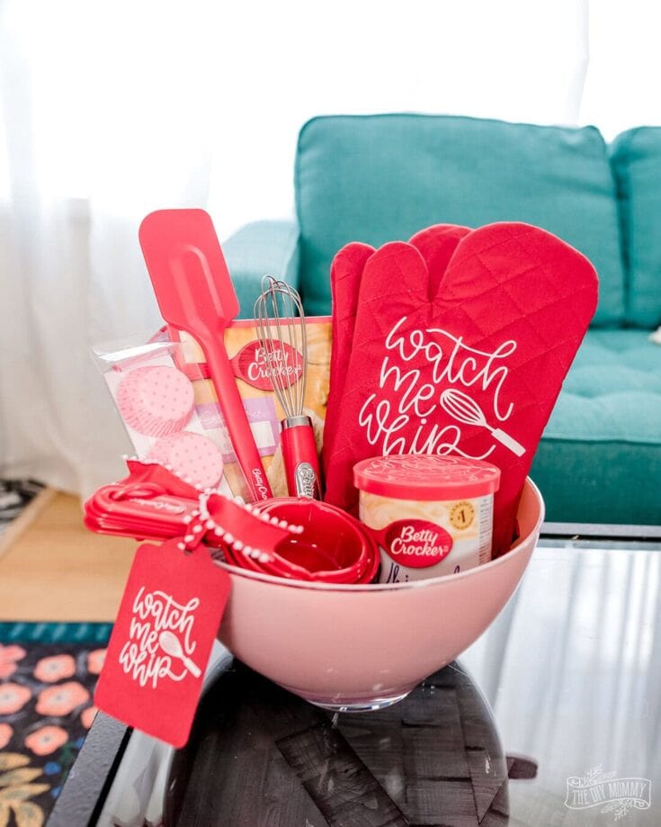 Red kitchen accessories and baking tools in a pink bowl, featuring "Watch Me Whip" oven mitt and labels, set on a black table with a teal sofa in the background, perfect for Valentine's Day or baking enthusiast themes.