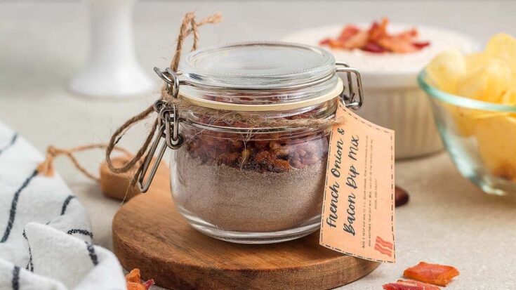 French Onion & Bacon Dip Mix in a glass jar on a round wooden board, with snacks and bowls in the background, perfect for appetizer gatherings.