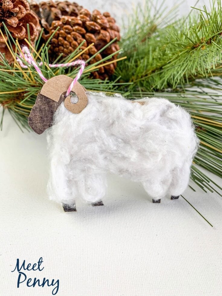 Soft white wool felt sheep ornament with stitched details, set against festive pine branches and pinecones, perfect for Christmas decor and holiday ornament crafts.
